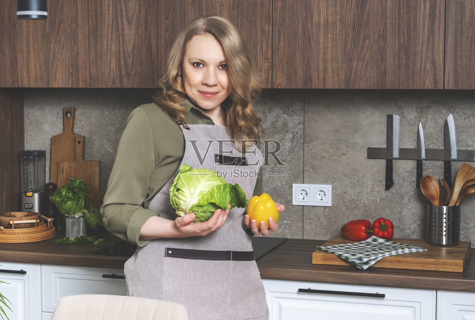 快乐微笑的金发中年女性家庭主妇手持新鲜的卷心菜和红椒。现代白色和木质厨房配备厨房电器和小工具。照片摄影图片