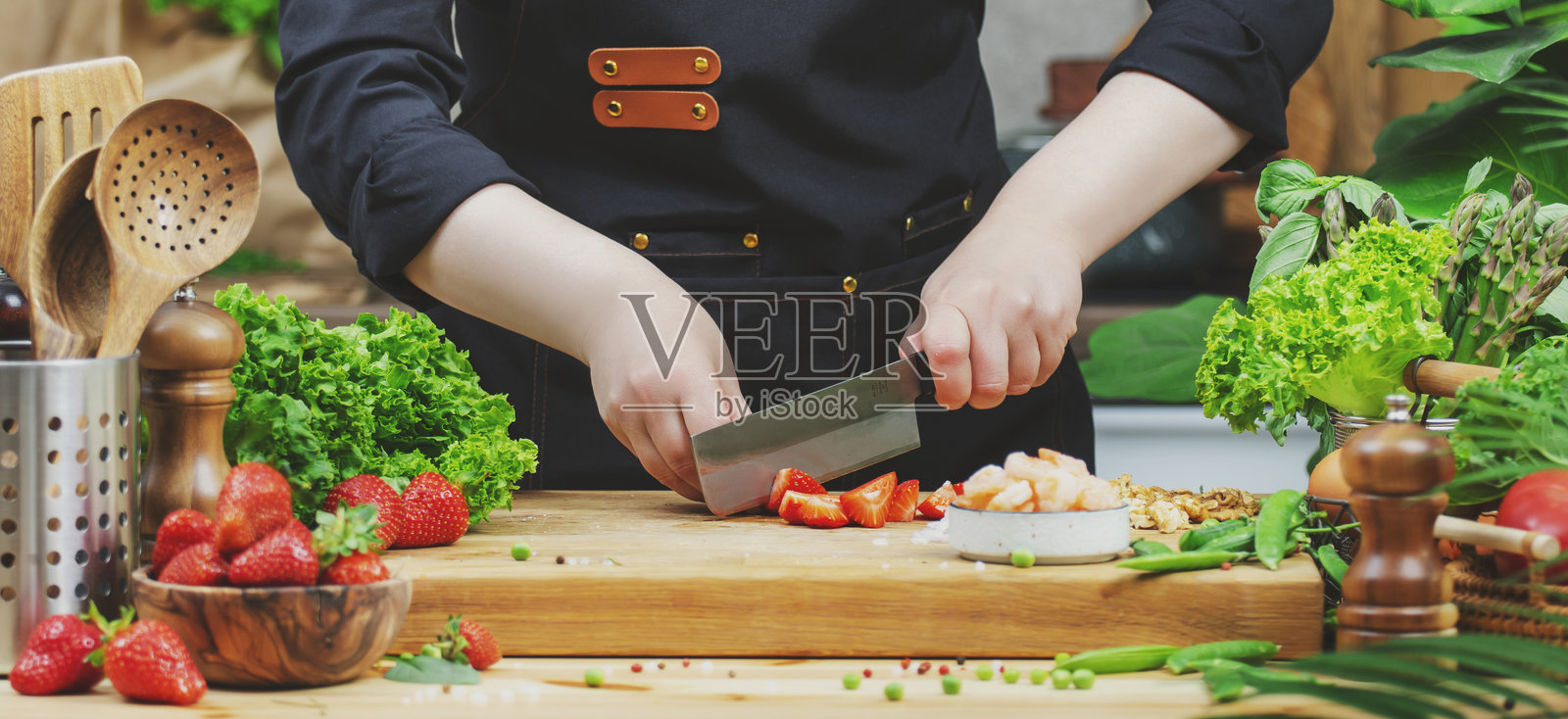 厨师在厨房烹饪食物。厨师切新鲜草莓，准备沙拉。木桌、厨房用具、蔬菜、香草和配料。健康的素食饮食、食谱、博客。照片摄影图片