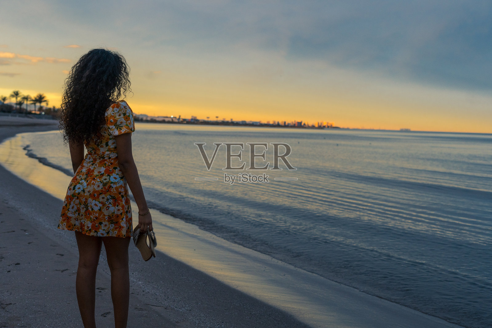 年轻女性在日落时分赤脚走在海滩上，手里拿着鞋子照片摄影图片