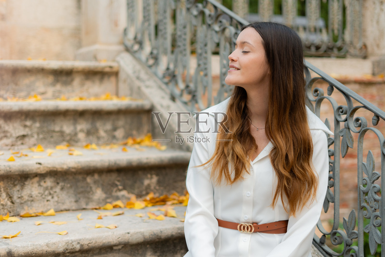 时尚的长发棕色女性穿着白衬衫，坐在覆盖着黄色秋叶的古老石阶上，闭着眼睛享受着清新的空气，旁边是一个锻铁栏杆。照片摄影图片
