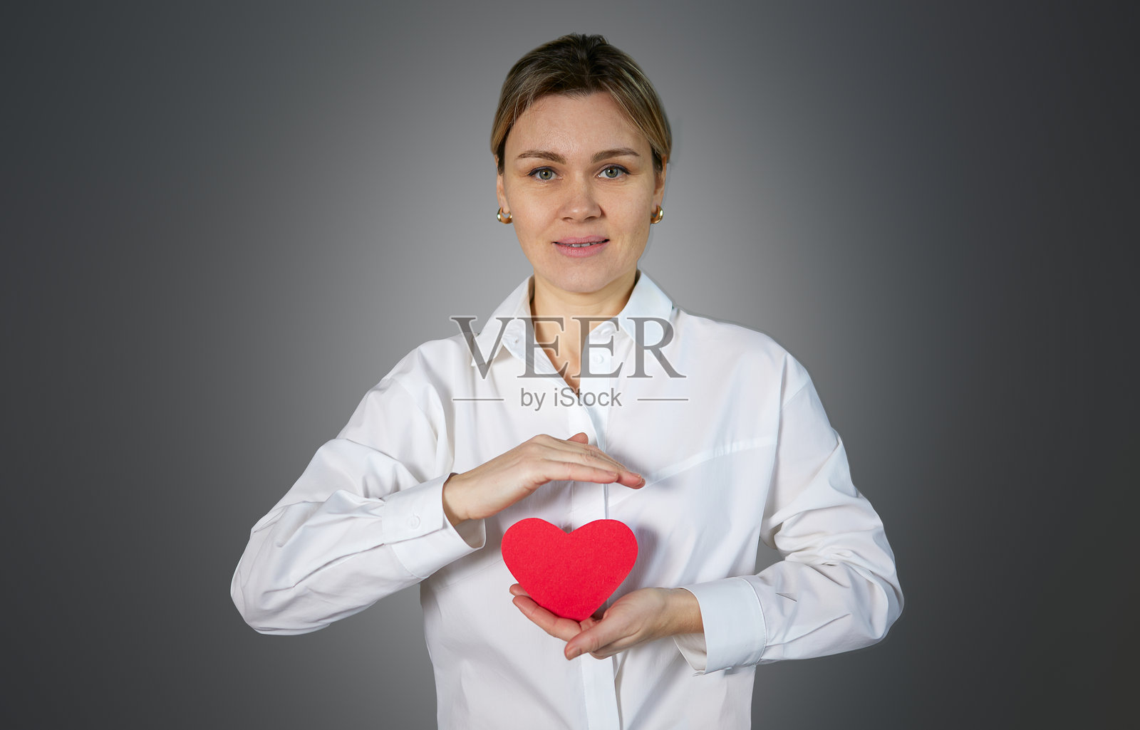 一位微笑的女性在灰色背景下双手捧着一个红色的心，象征保护、庇护和珍惜。照片摄影图片