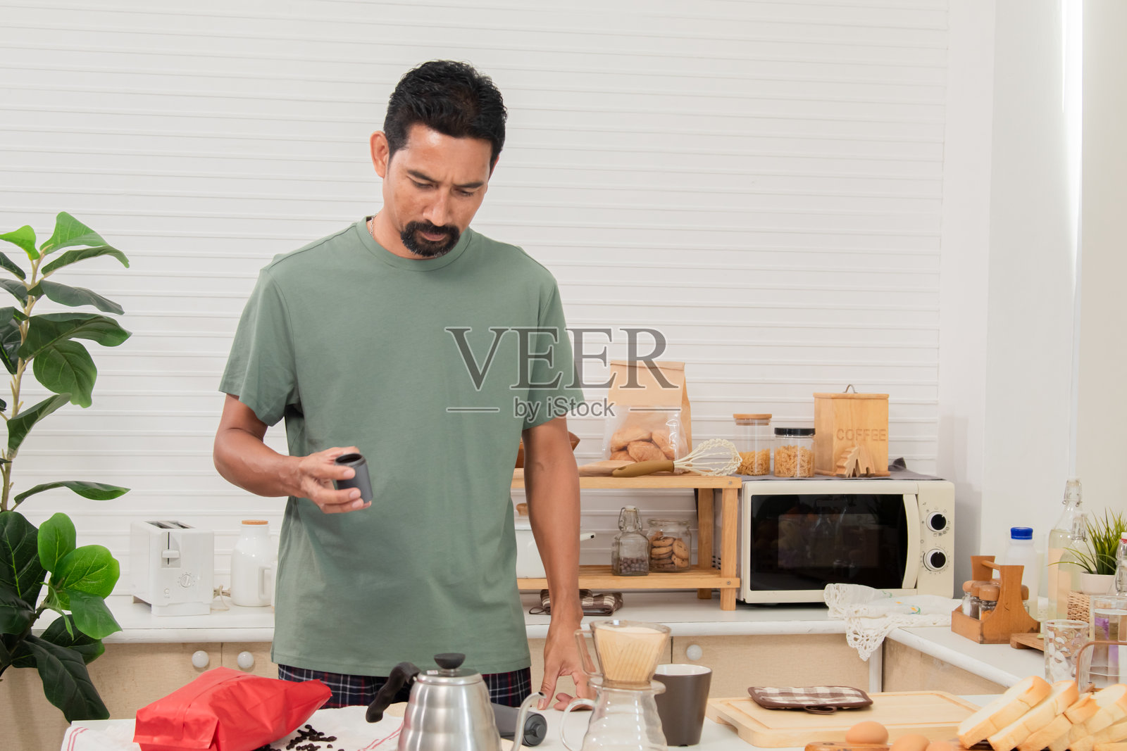 中年有胡子的亚洲男子在家中使用手摇咖啡磨豆机研磨咖啡豆，将其磨成粉，以便早晨饮用。健康生活方式概念。照片摄影图片