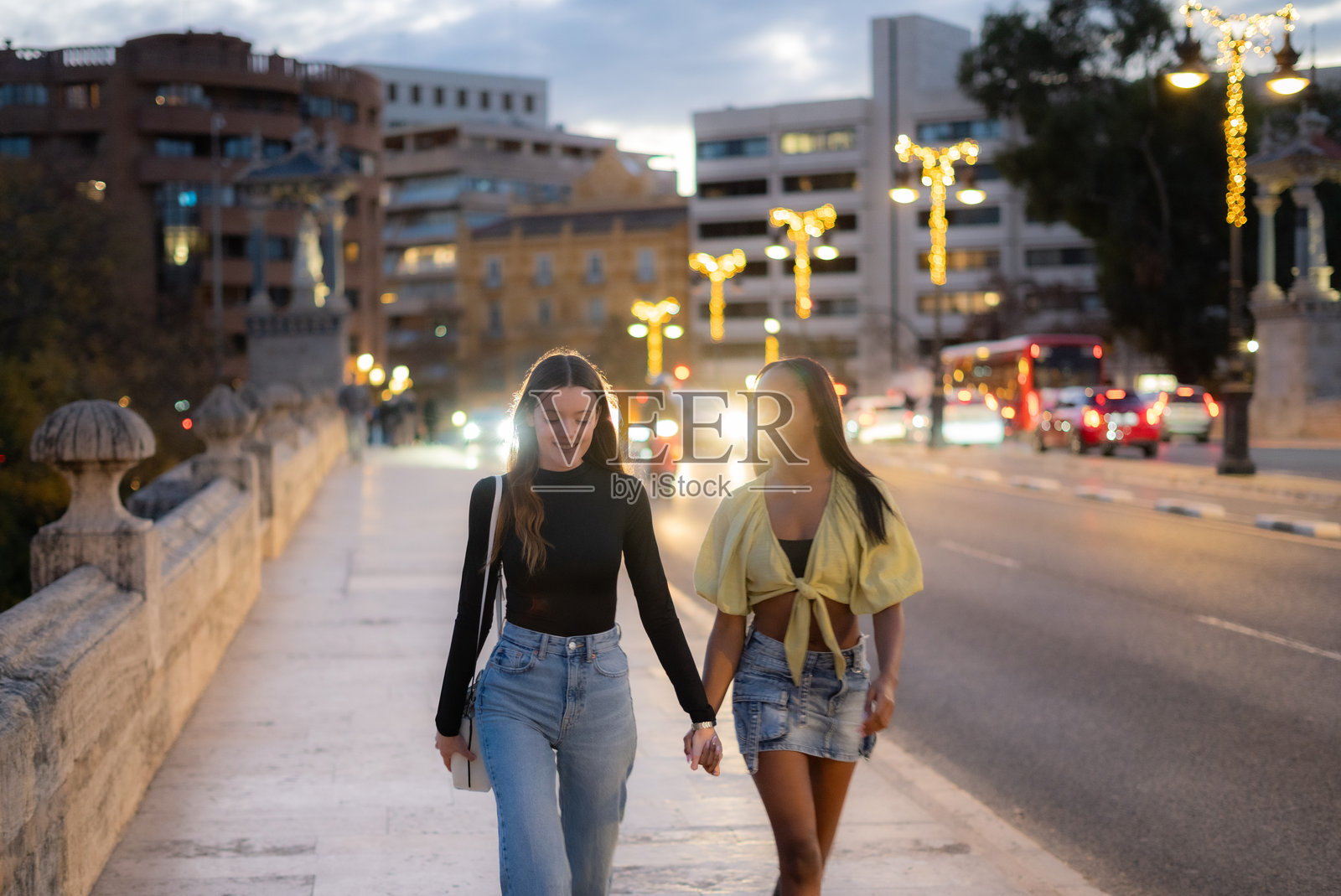 两位年轻女性游客在西班牙瓦伦西亚的桥上散步，黄昏时分，她们手牵手，享受着城市的灯光和夜晚的氛围。照片摄影图片