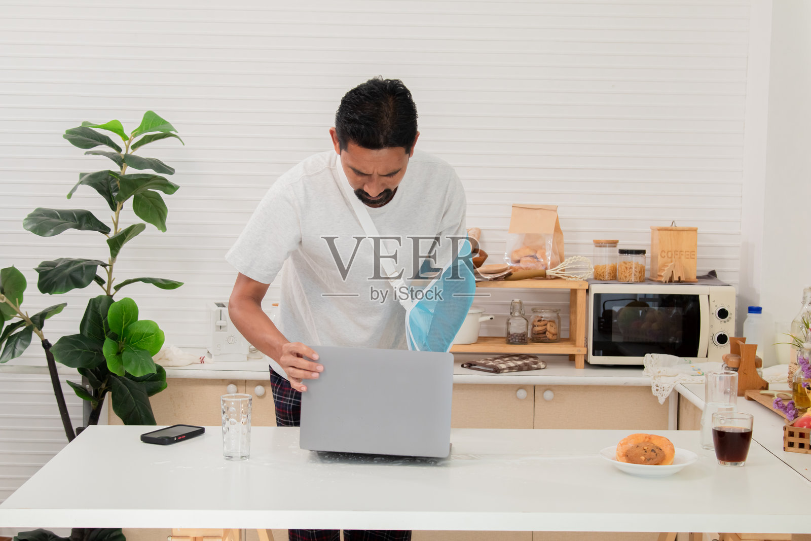 中年男子因右臂受伤而带着吊带在家中病假，早晨在工作。受伤的商人专注地看着笔记本电脑并使用智能手机。照片摄影图片