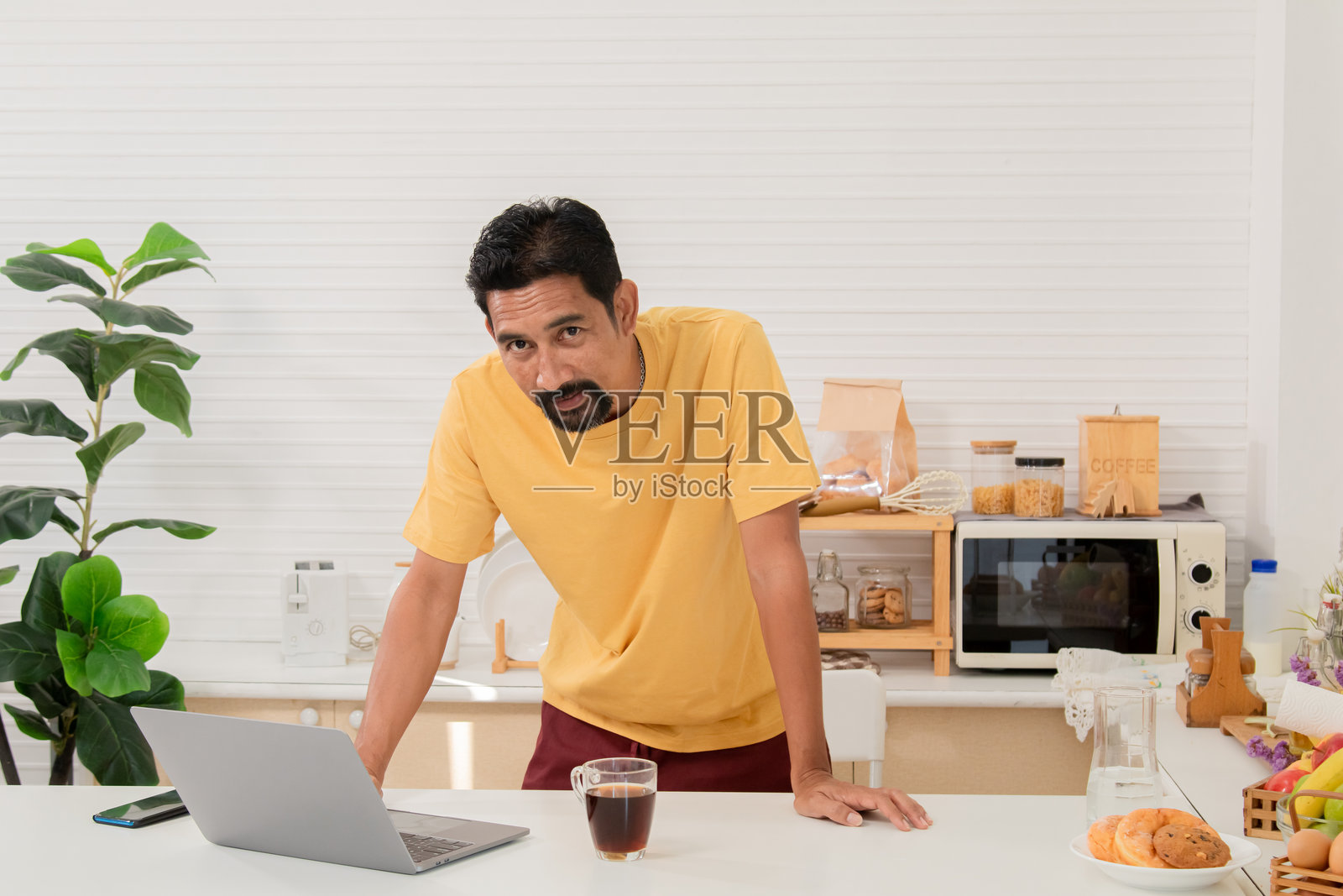 中年有胡子的亚洲男子拿着笔记本电脑走进厨房，准备为早餐拿些牛奶。自由职业者在家工作，早晨阅读新闻。成功的男性企业家在家办公。照片摄影图片