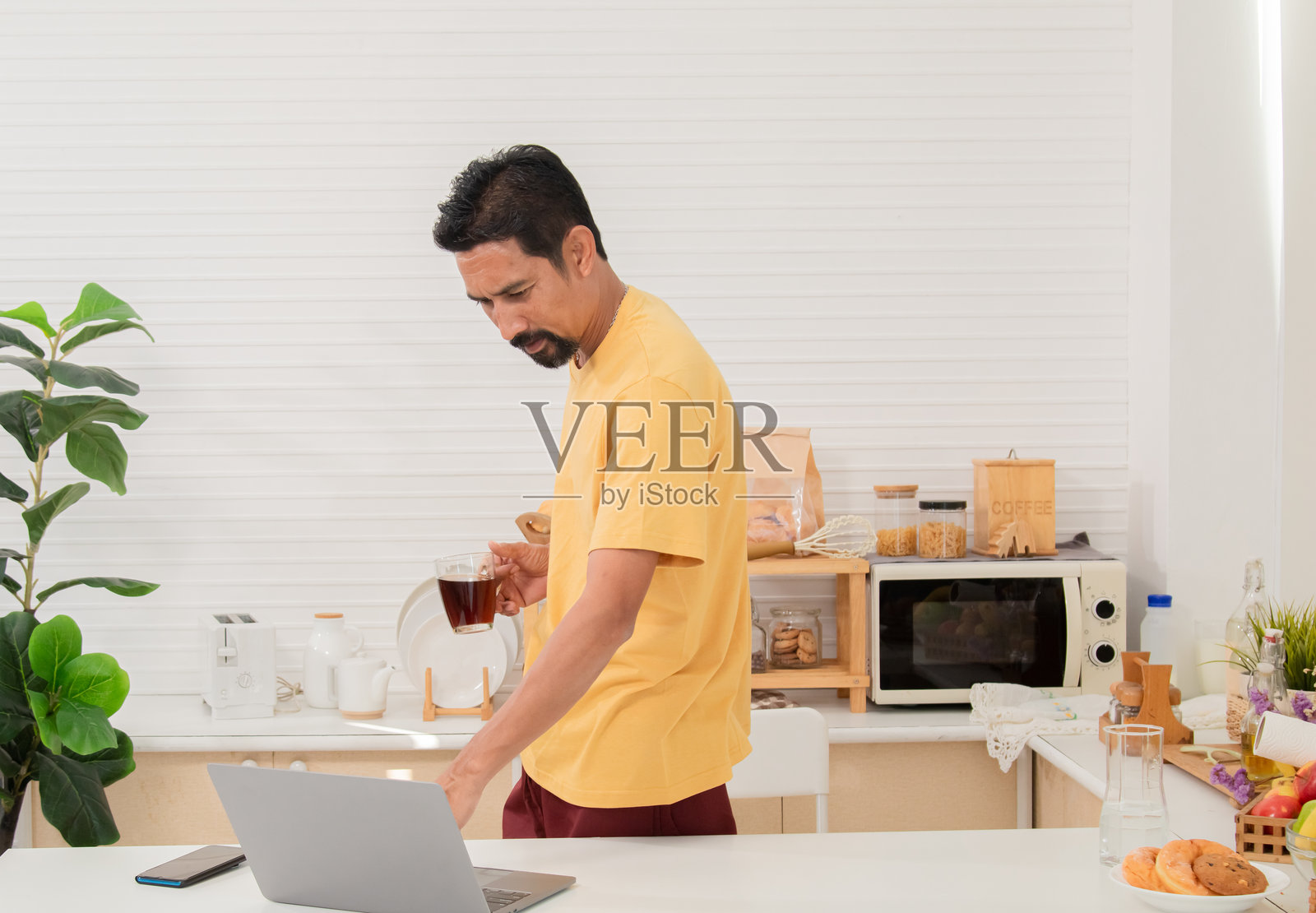 中年有胡子的亚洲男子拿着笔记本电脑走进厨房，准备为早餐拿些牛奶。自由职业者在家工作，早晨阅读新闻。成功的男性企业家在家办公。照片摄影图片