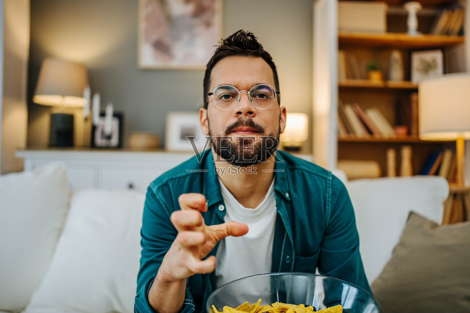 年轻男子坐在沙发上看电视，边吃碗里的玉米片。照片摄影图片
