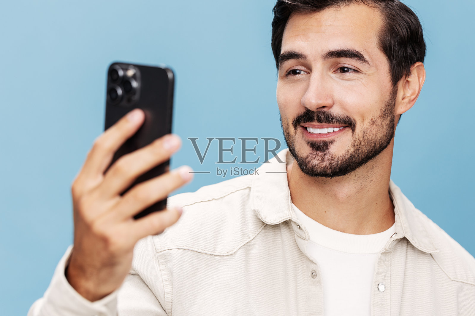 棕发男子特写，惊讶地看着手机屏幕，嘴巴微张，露出快乐的表情，背景为蓝色，穿着白色T恤和牛仔裤，留有空白空间。照片摄影图片