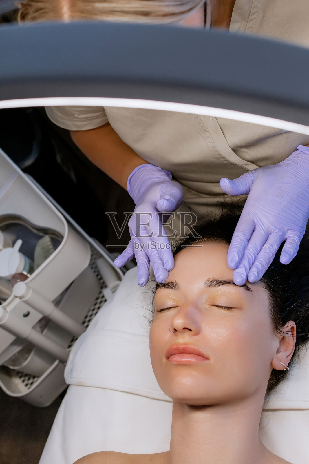 专业美容师戴着紫色乳胶手套，在现代美容院水疗诊所为躺在治疗台上的年轻女性客户进行面部皮肤护理。照片摄影图片
