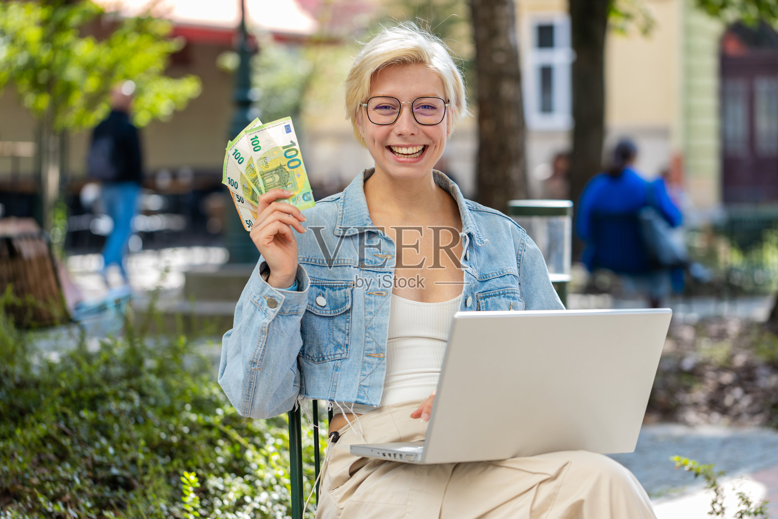 快乐富有的年轻女性在城市中使用笔记本电脑，胜利地手握欧元现金。照片摄影图片