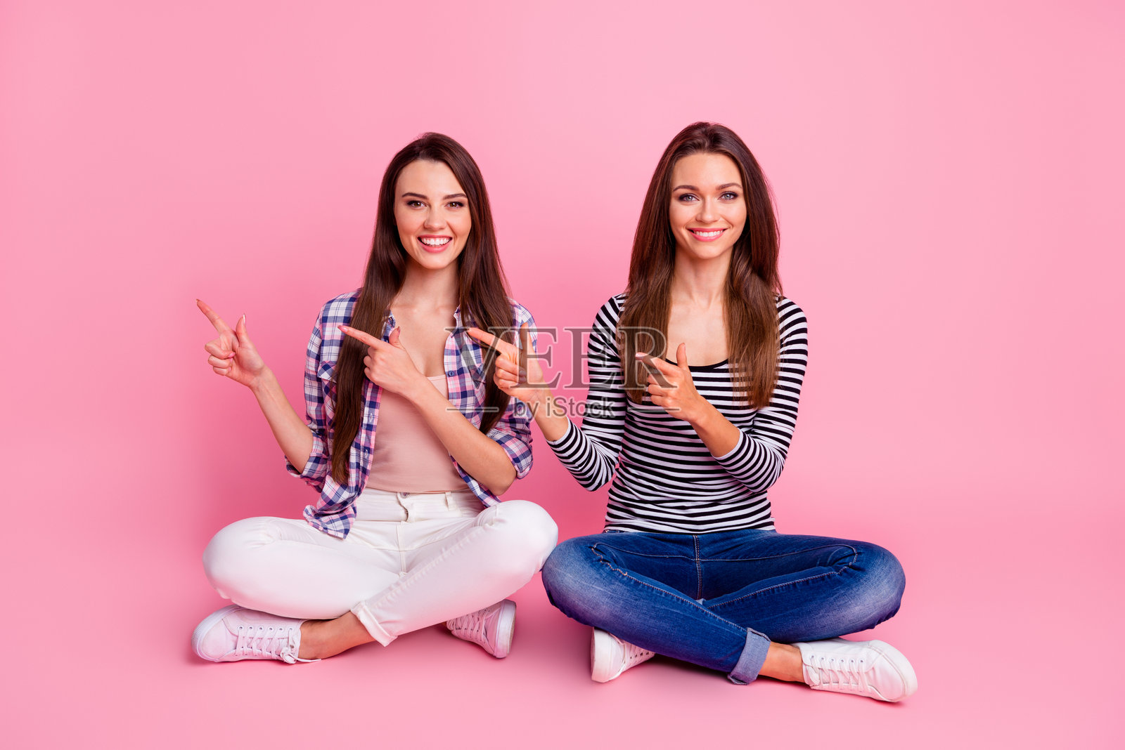 两位漂亮开朗的人坐在地板上，手指指向空旷的空间，背景为粉色。照片摄影图片