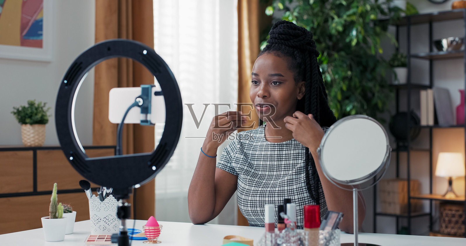 一位非裔美国女性主持直播美容课程，分享她的化妆流程，向粉丝问好，坐在化妆台旁，配有环形灯和三脚架的智能手机。照片摄影图片