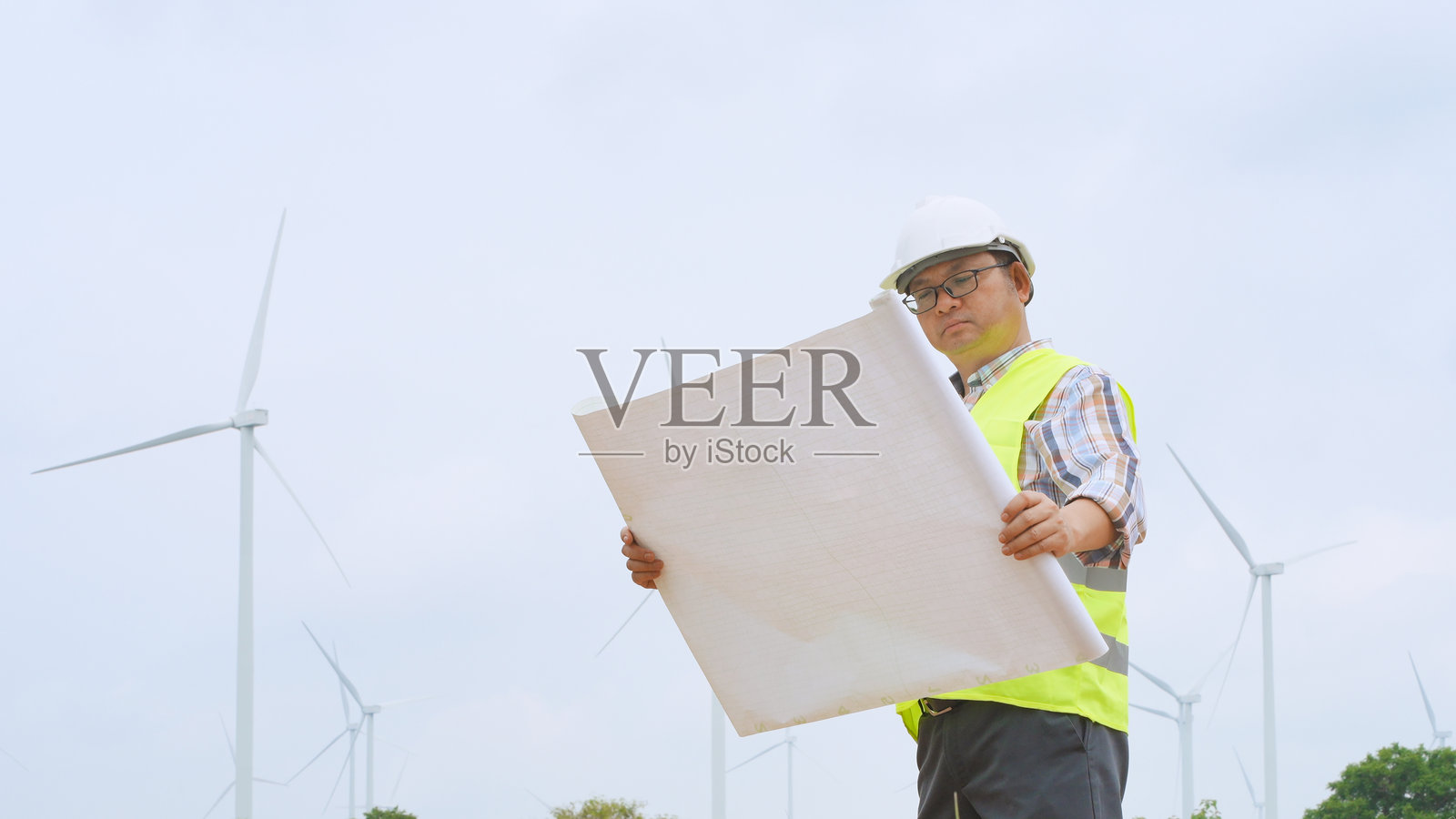 工程师在明亮的天空下查看可再生能源现场的风力涡轮机，手中拿着蓝图。照片摄影图片