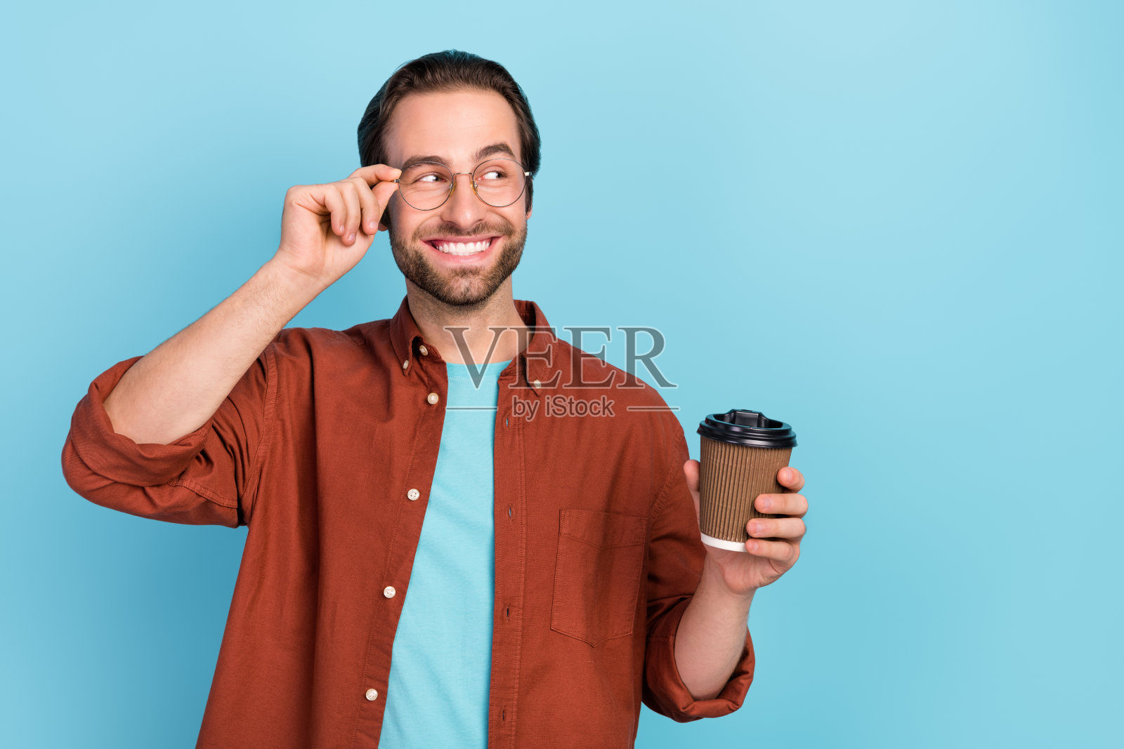 乐观的棕发千禧一代男士手持咖啡，穿着棕色衬衫，戴着眼镜，背景为蓝色。照片摄影图片
