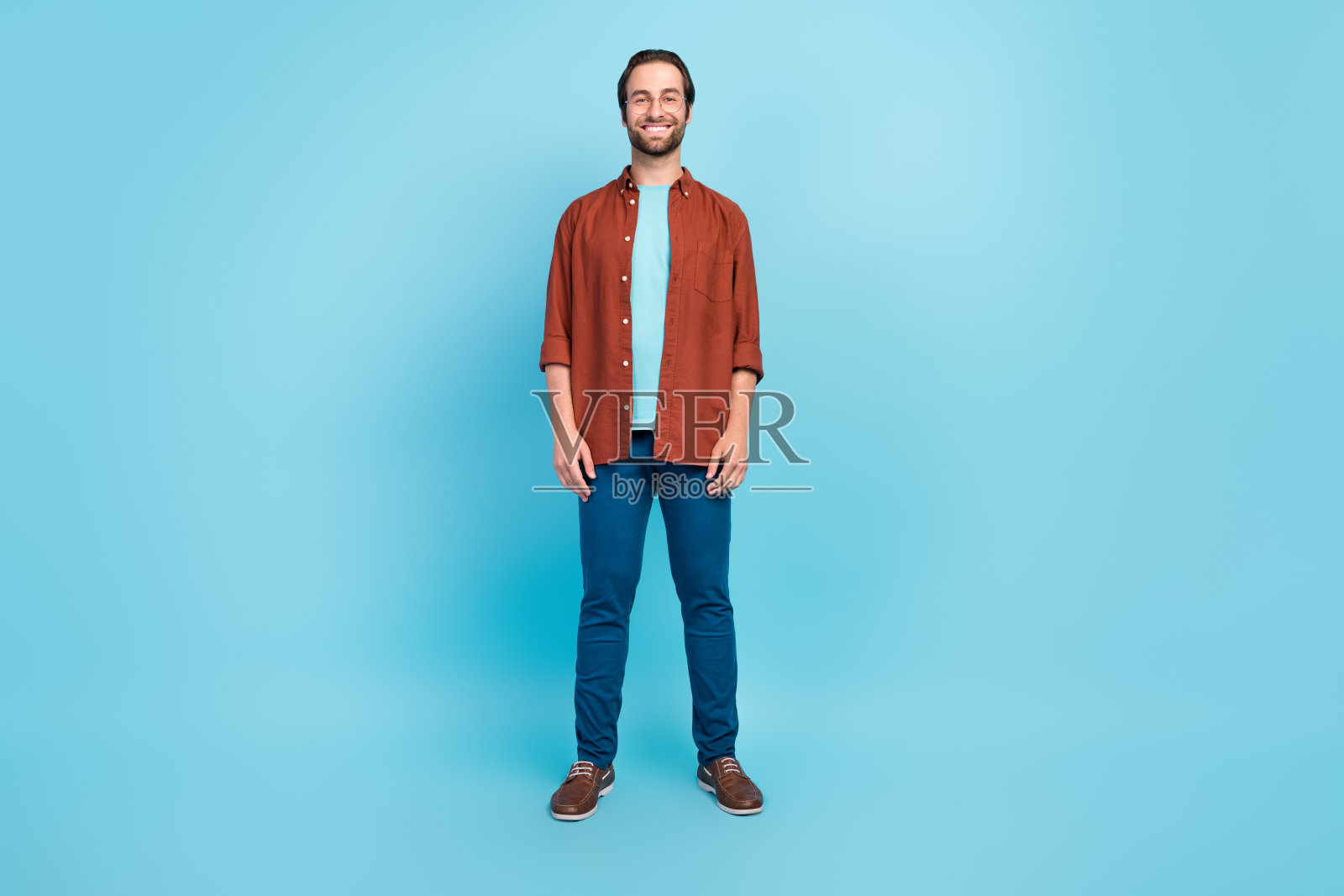 全身照：一位穿着衬衫、长裤和运动鞋的帅气棕发千禧一代男士，戴着眼镜，背景为蓝色。照片摄影图片