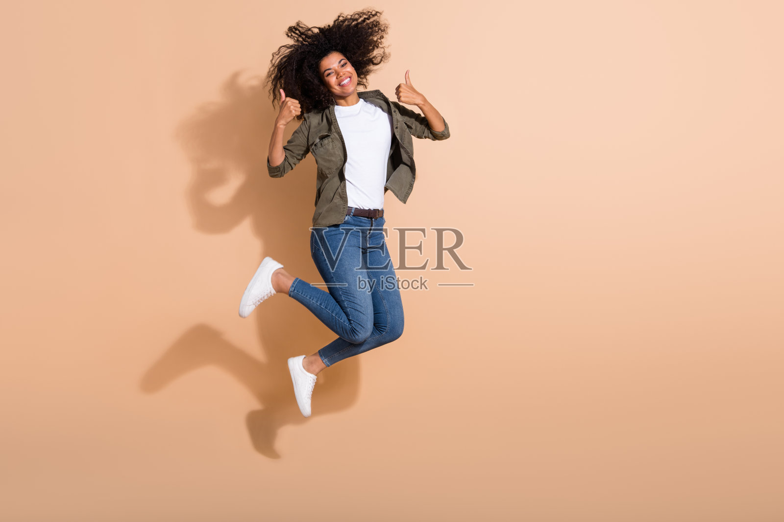 全身长视图的迷人快乐女孩跳跃并竖起大拇指，背景为米色柔和色调。照片摄影图片