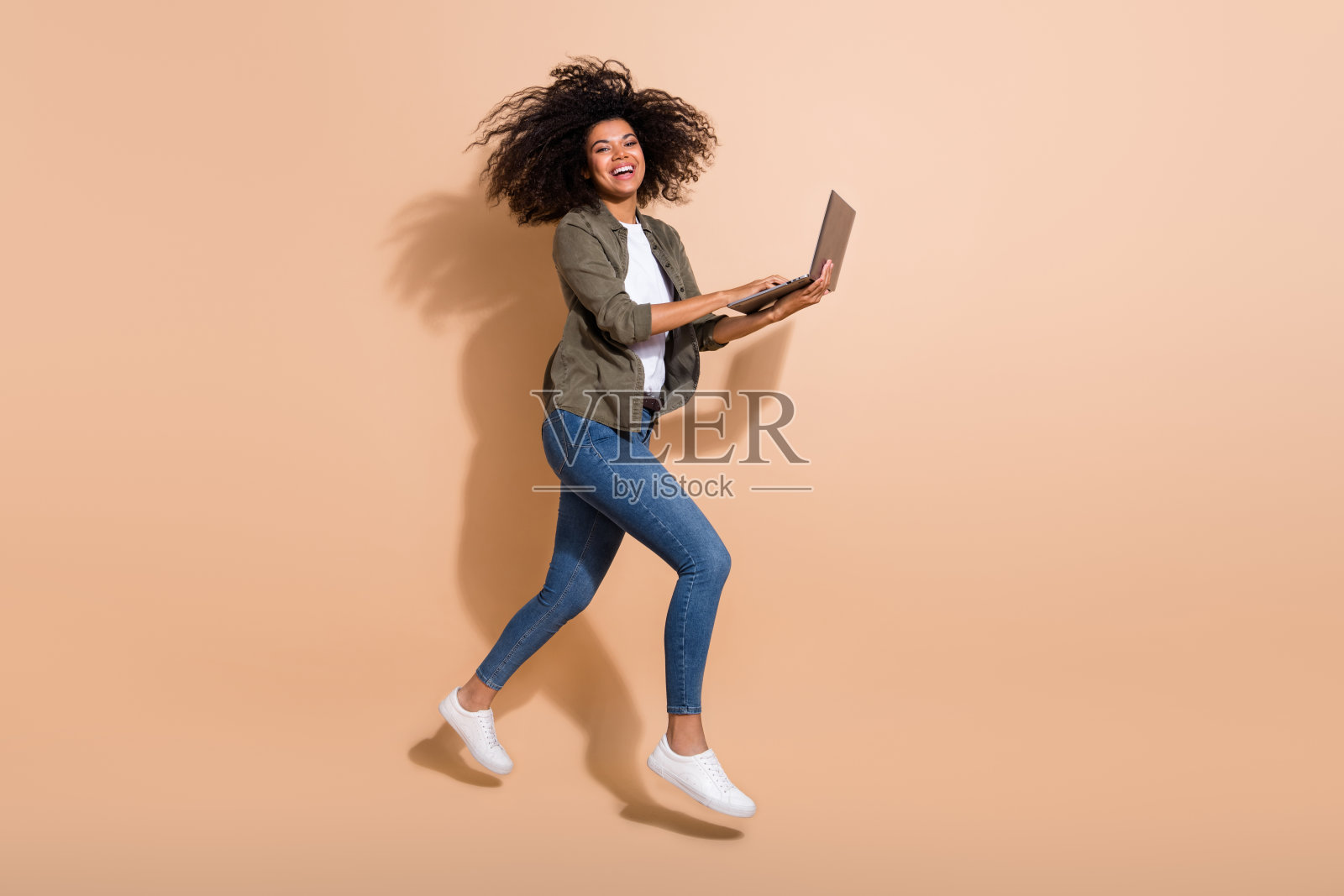 全身身材的迷人快乐女孩在米色柔和背景下跳跃着使用笔记本电脑，享受乐趣。照片摄影图片