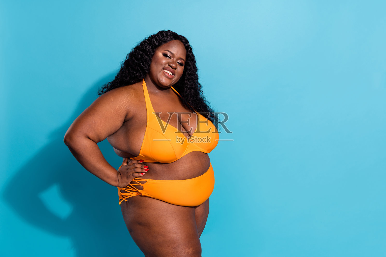胖胖的女士穿着比基尼，双手放在腰间，站在蓝色背景下的空旷空间中，爱着自己的身体。照片摄影图片