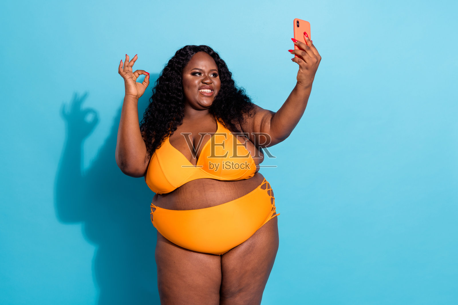 时尚前卫的胖女人在明亮的蓝色背景下自拍，做出“好”的手势，分享生活。照片摄影图片