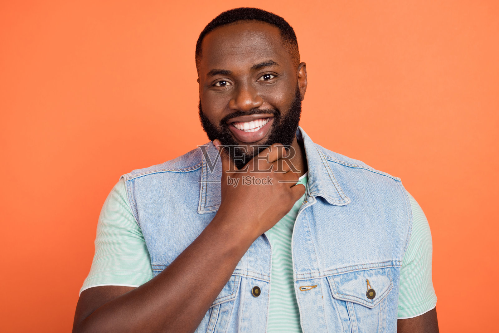 穿着牛仔背心的聪明可爱的非洲裔男孩，手臂托着下巴，微笑着，背景为橙色。照片摄影图片