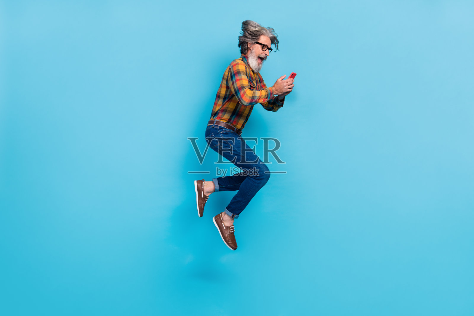 兴奋的退休男士穿着格子衬衫，戴着眼镜，跳得很高，手里拿着现代设备，背景为蓝色。照片摄影图片