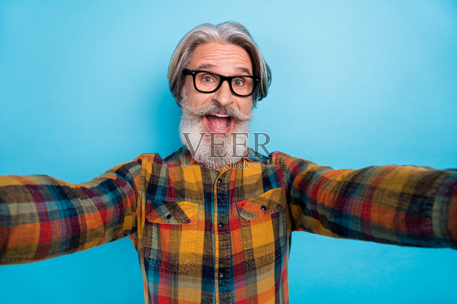 穿着格子衬衫、戴着眼镜的时尚退休男士在蓝色背景下张嘴自拍，显得十分惊讶。照片摄影图片