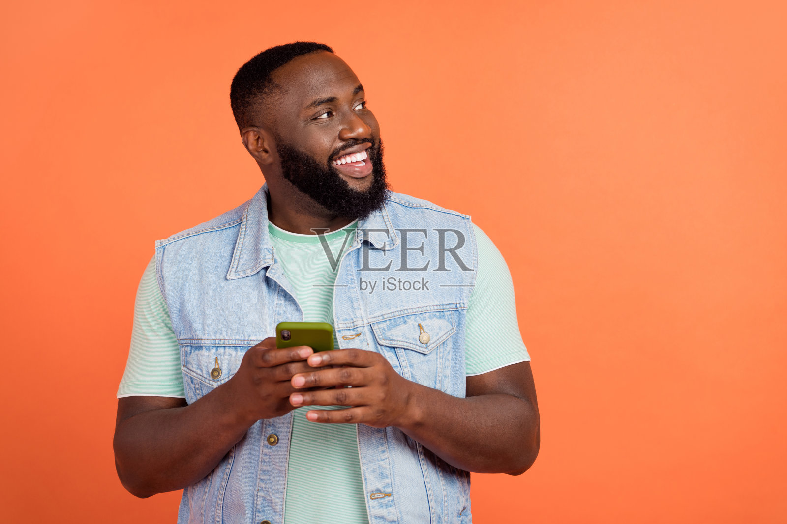 穿着牛仔裤和马甲的梦幻男孩在橙色背景下打字，目光空洞。照片摄影图片