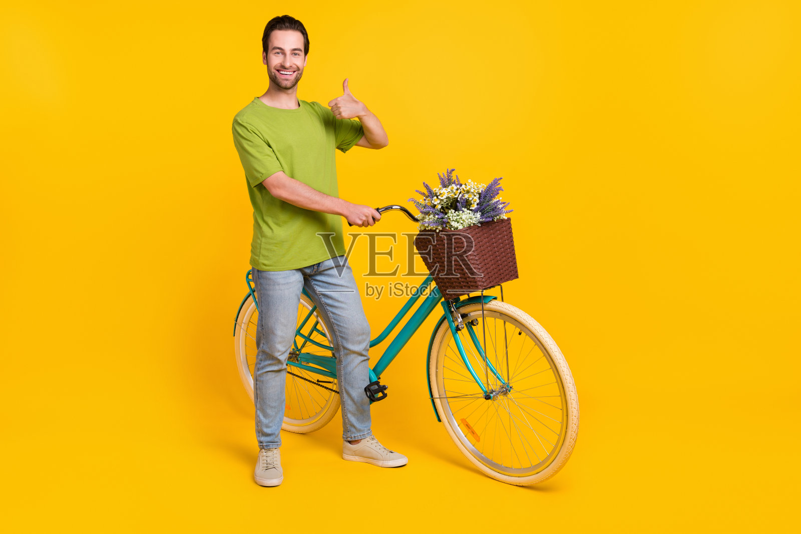 全身照，酷炫的棕发千禧一代男孩骑自行车，竖起大拇指，穿着T恤、牛仔裤和鞋子，背景为黄色。照片摄影图片