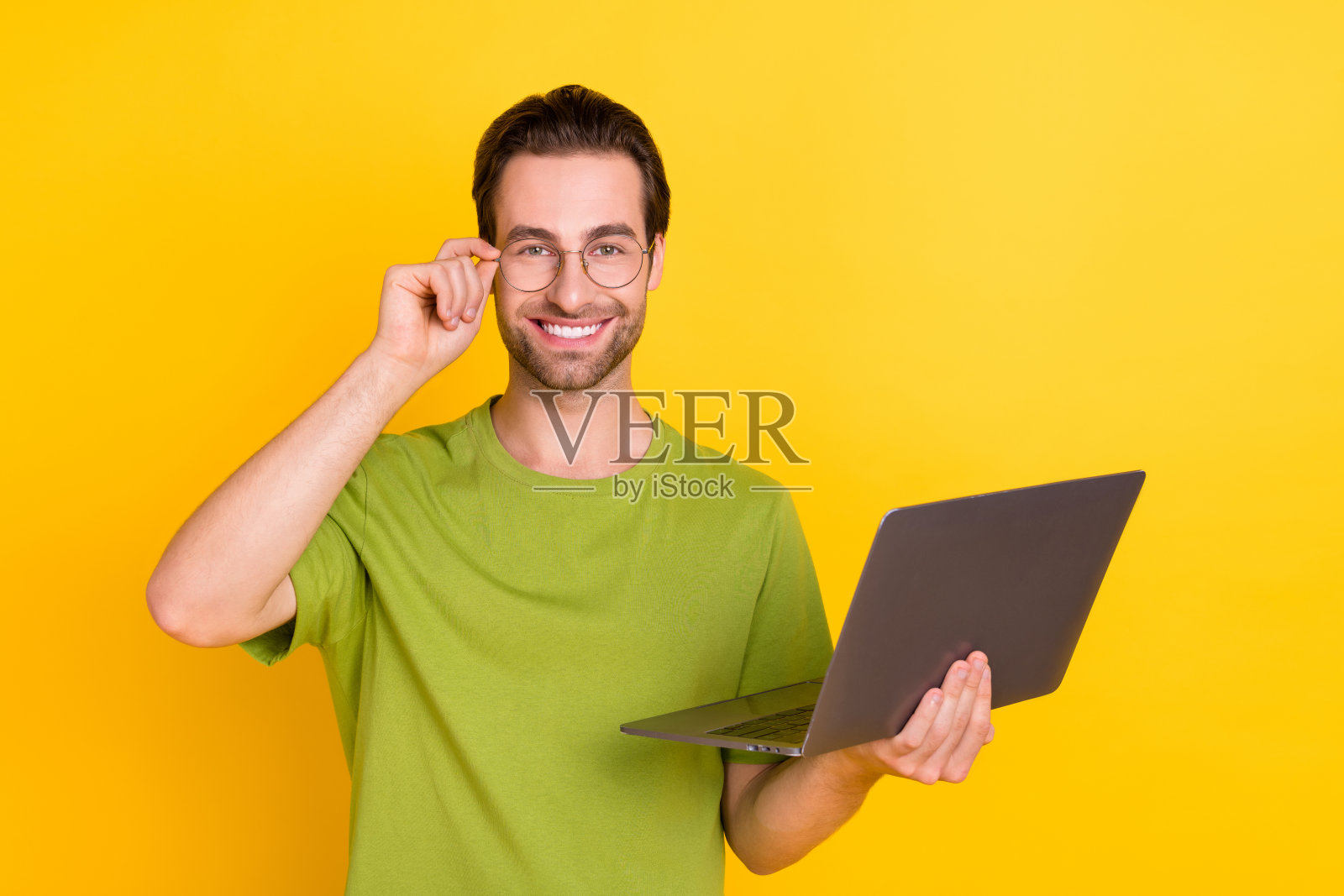 可爱的胡须千禧男士戴着眼镜，穿着绿色T恤，手持笔记本电脑，背景为黄色。照片摄影图片
