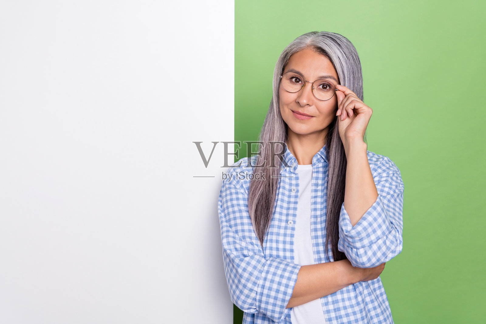一位白发苍苍的思考老年女性，戴着眼镜，穿着格子衬衫，站在绿色背景前的海报旁。照片摄影图片