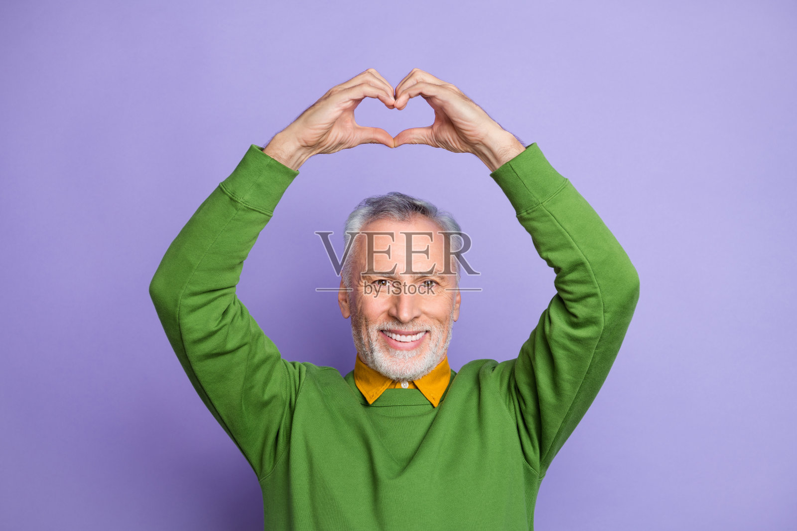 穿着绿色毛衣的迷人退休老人双手举起，心形手势，背景为紫色。照片摄影图片