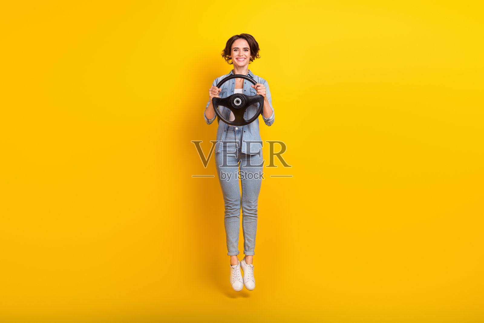 年轻女性在黄色背景下充满活力地跳起，手握方向盘，享受假期的全身照片照片摄影图片