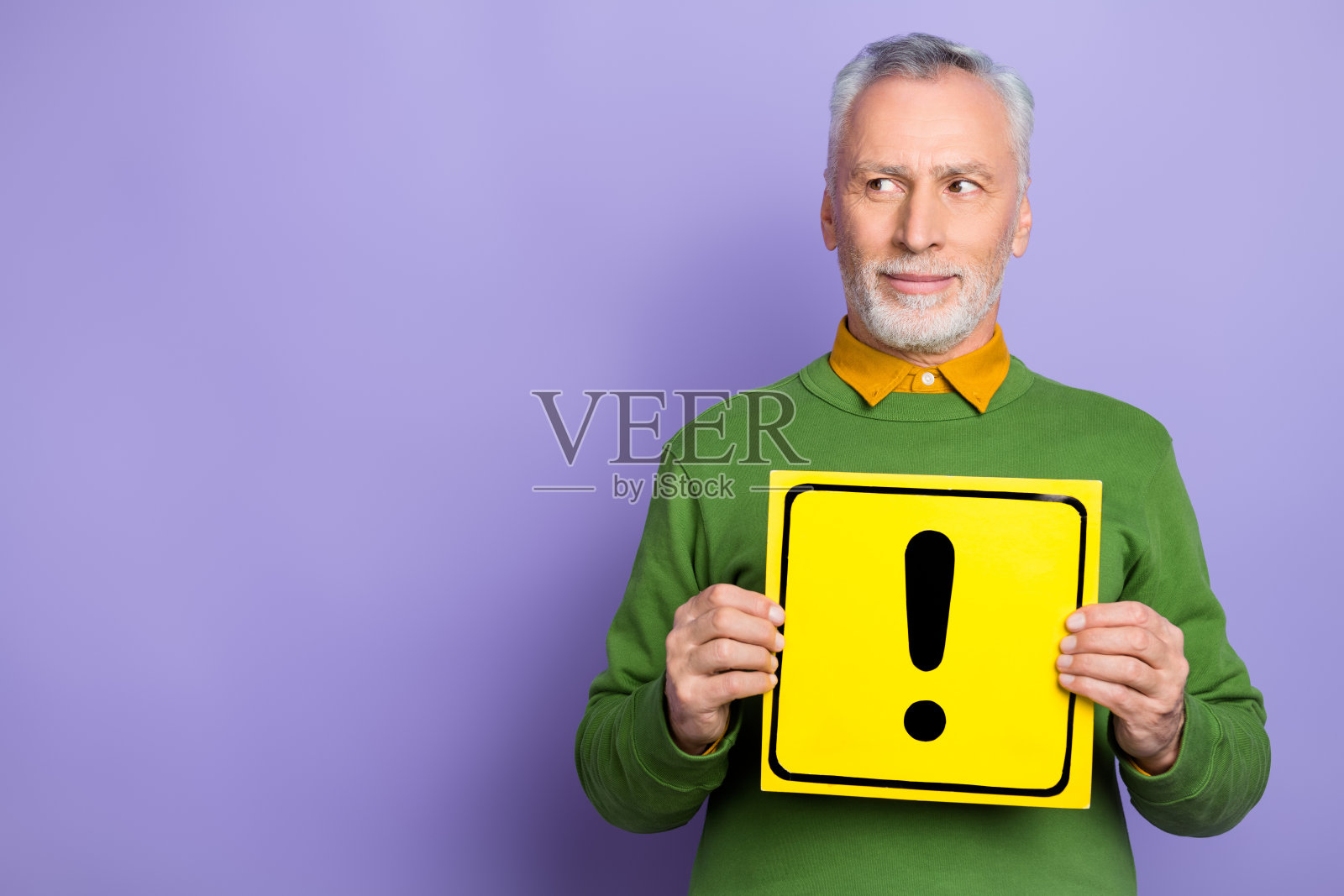穿着绿色毛衣的梦幻老年男子手持危险卡，空白区域，背景为紫色。照片摄影图片