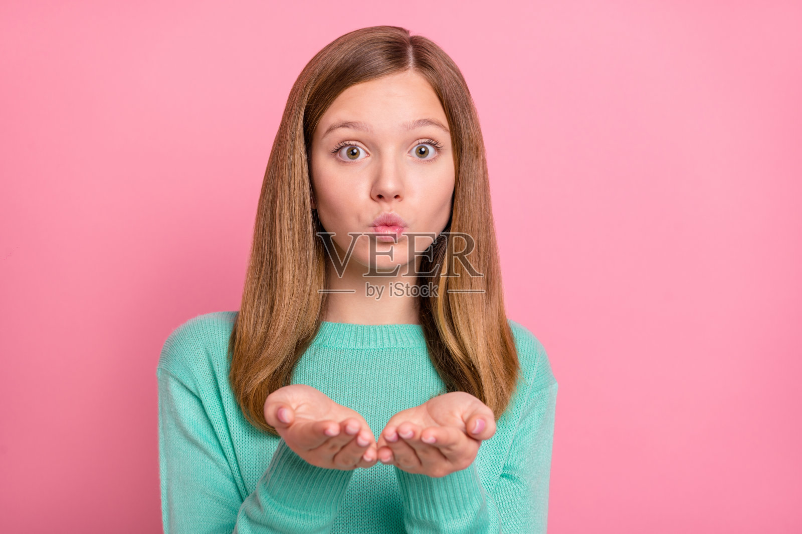 可爱的女学生穿着青绿色毛衣，向镜头送出飞吻，背景为粉色。照片摄影图片