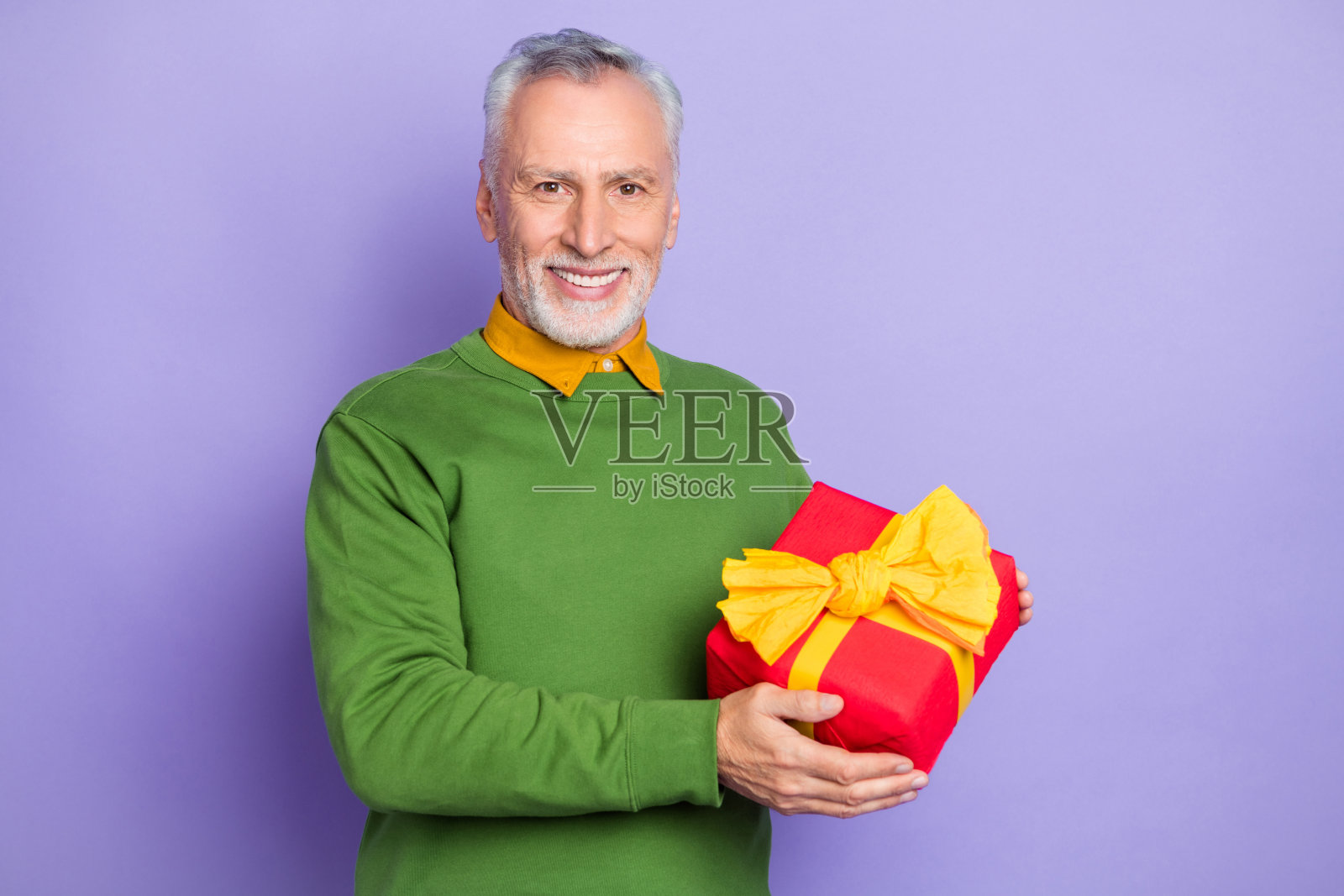 可爱的退休男士穿着绿色毛衣，手里拿着礼物包，背景为紫色。照片摄影图片