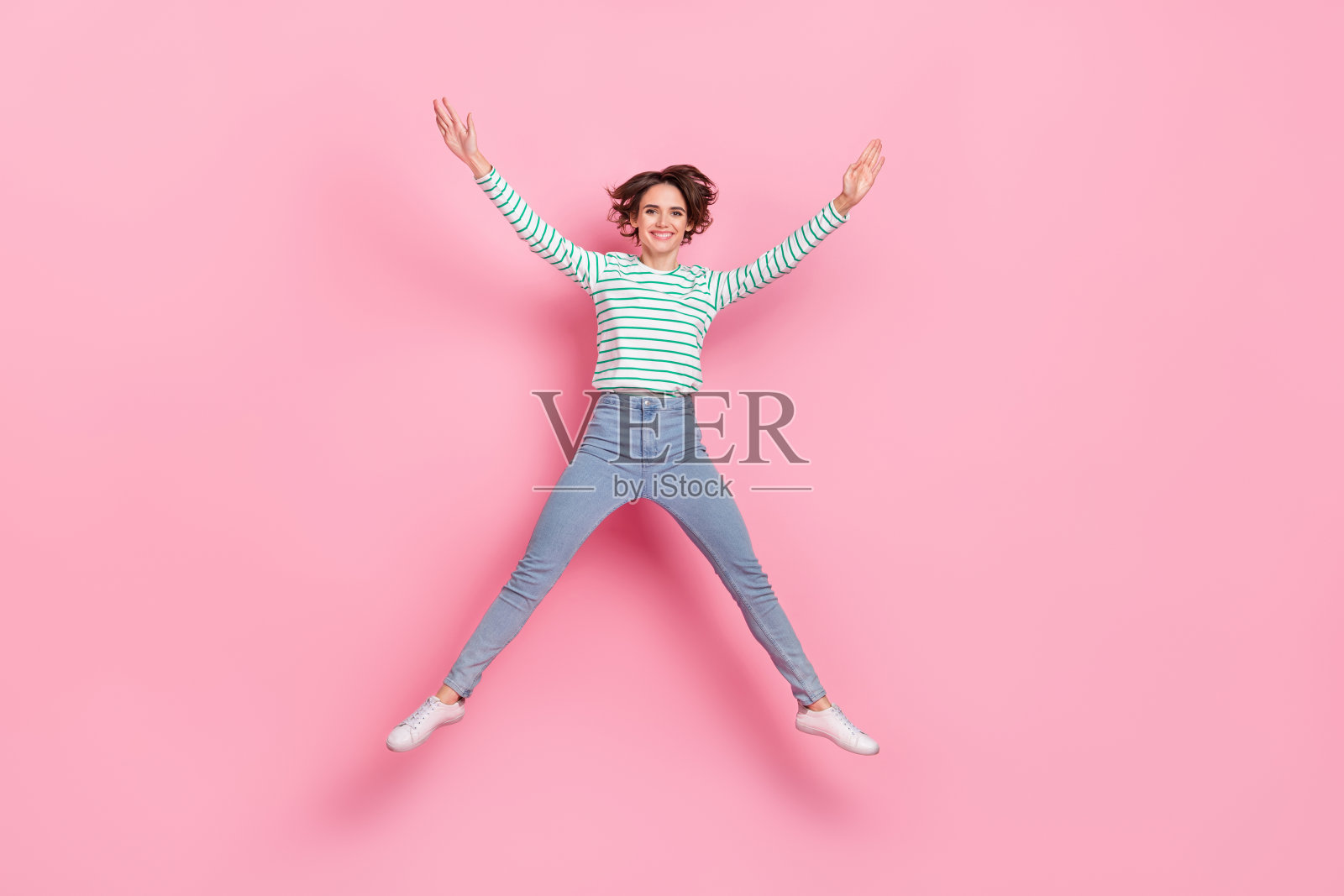 全身照，令人印象深刻的千禧一代短发女士跳跃，穿着衬衫、牛仔裤和鞋子，背景为粉色。照片摄影图片