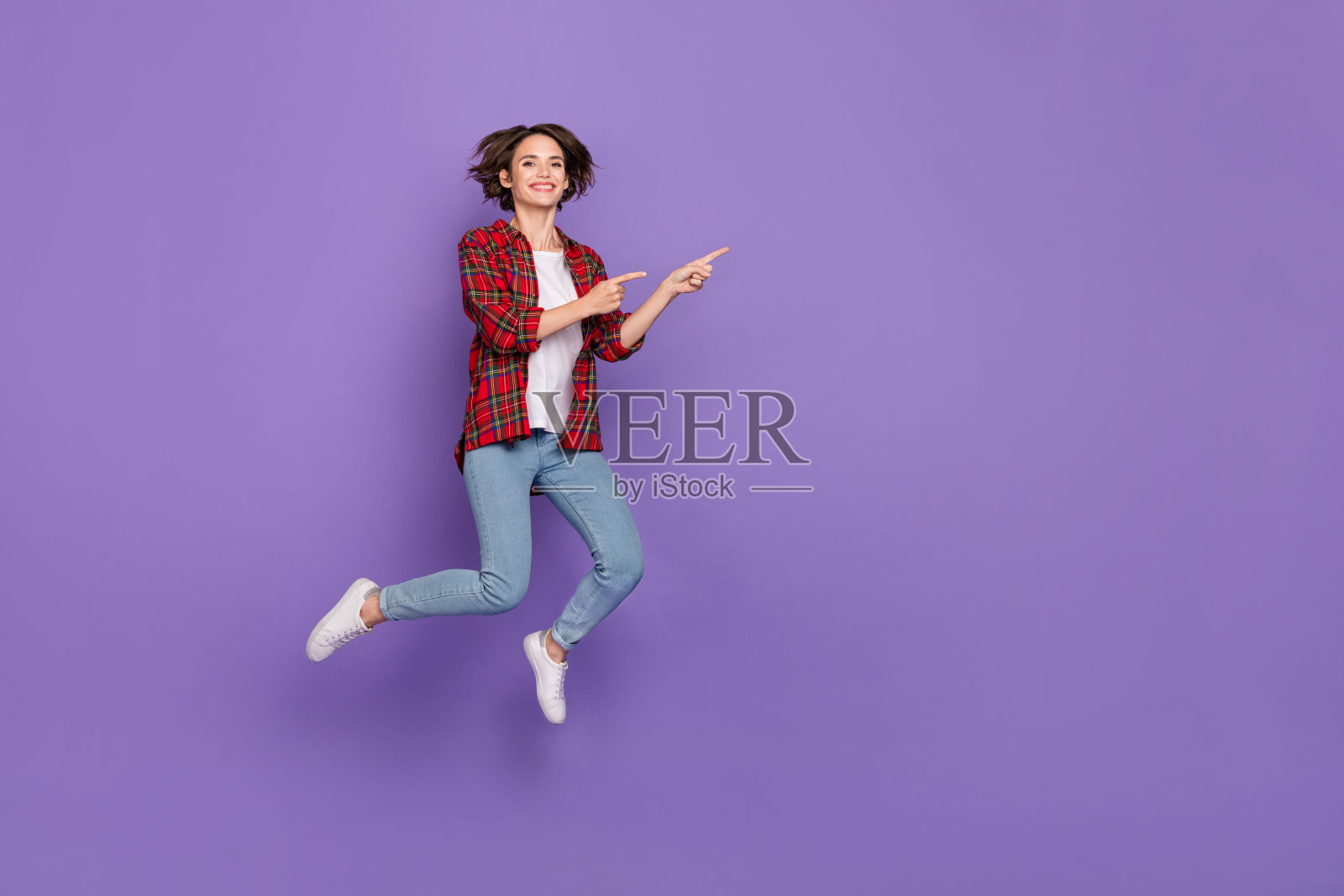 全身照，搞笑的年轻棕发女士穿着衬衫、牛仔裤和运动鞋，跳跃着，背景为紫色。照片摄影图片
