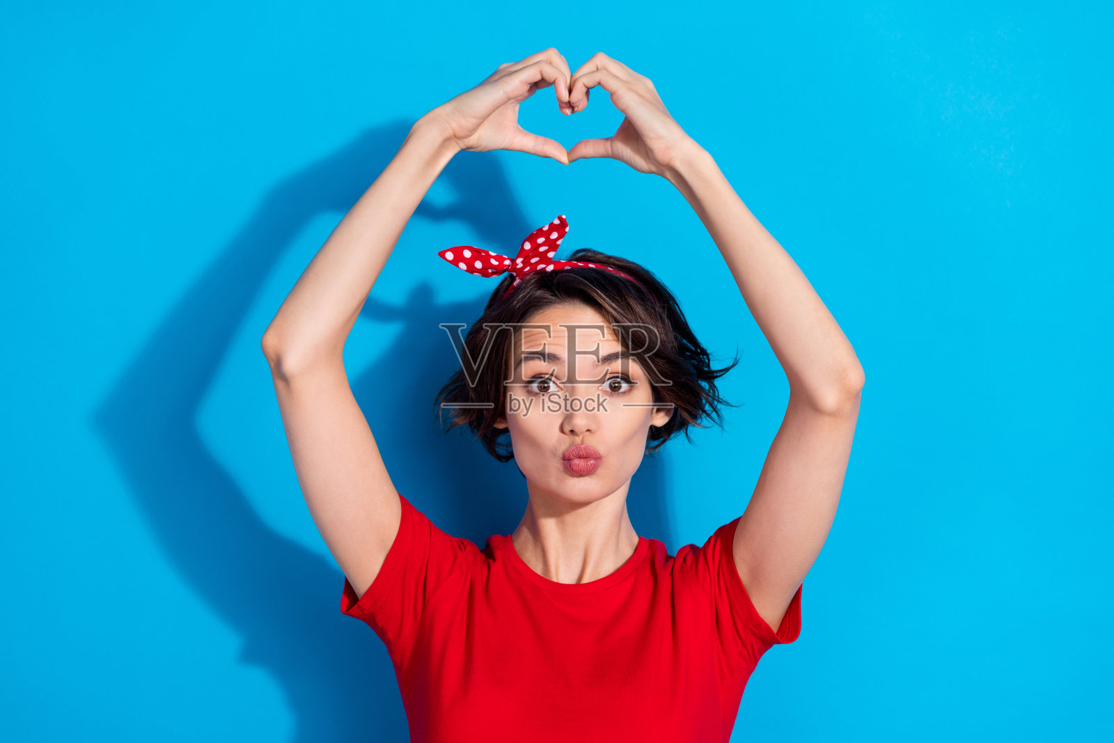 甜美的年轻棕发女孩穿着红色T恤，手捧心形物品，背景为蓝色。照片摄影图片