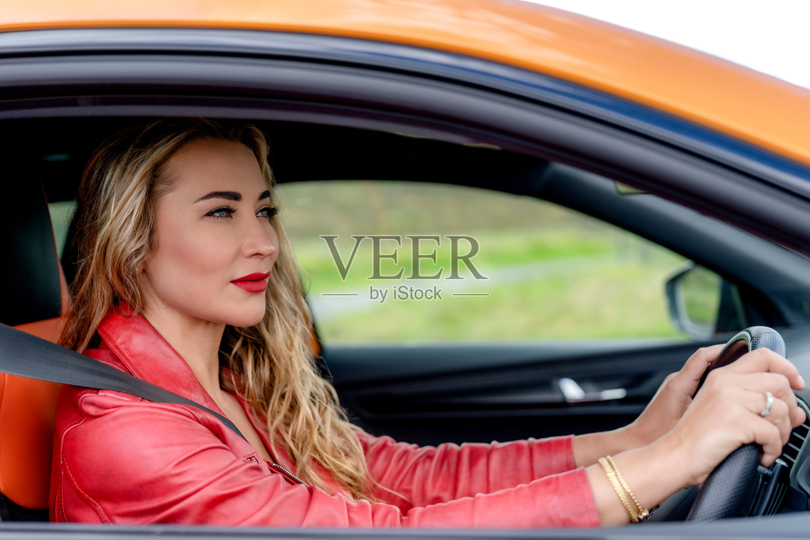 一位女性在阳光明媚的日子里，驾驶着一辆橙色汽车，沿着风景如画的道路行驶，背景是生机勃勃的绿色植物。照片摄影图片