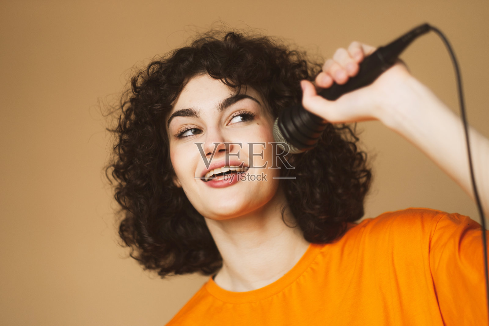 快乐的卷发年轻女性对着麦克风唱歌并跳舞。照片摄影图片