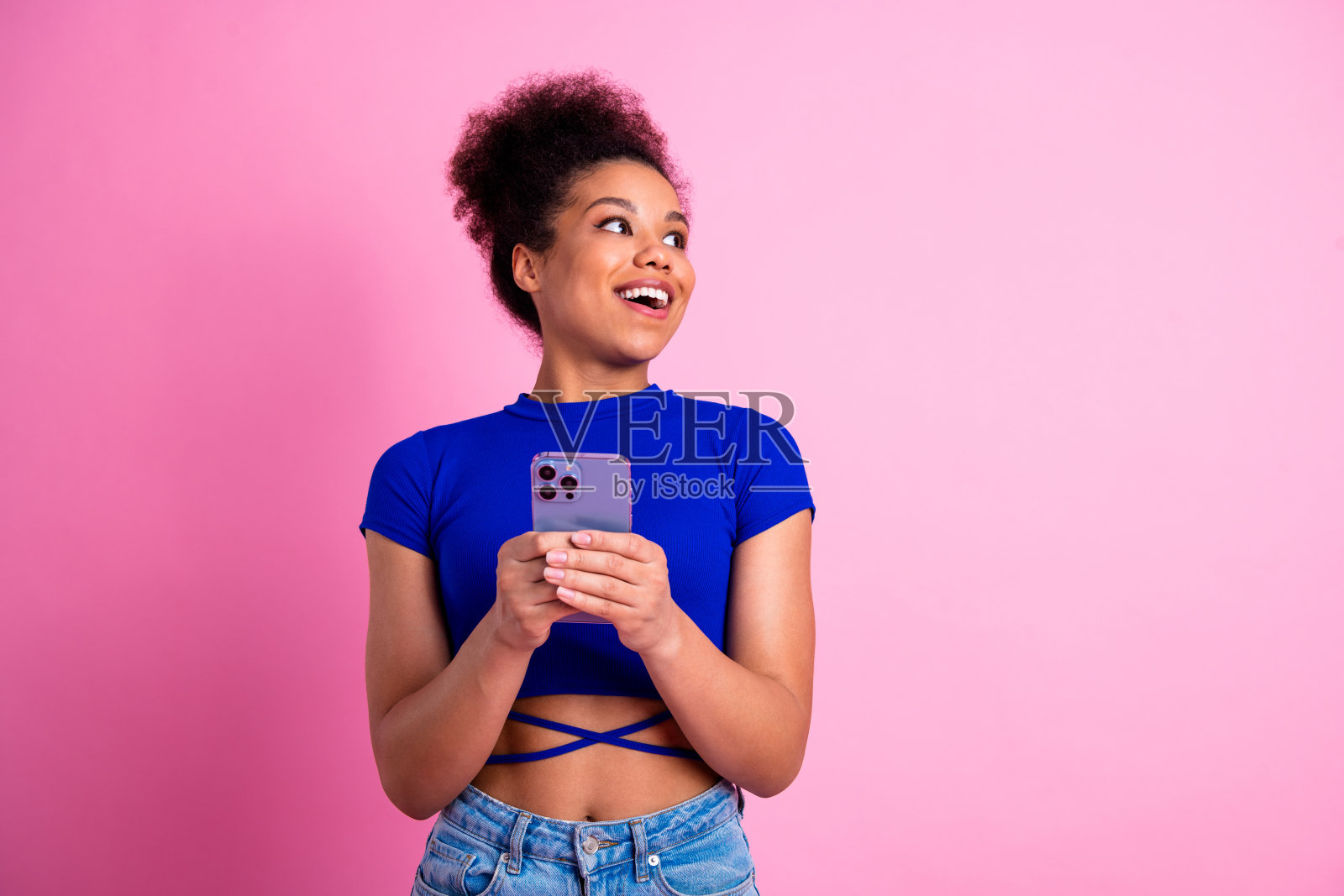 年轻女性手持智能手机，面带愉悦和互动的表情，背景为粉色，穿着时尚的蓝色上衣。照片摄影图片