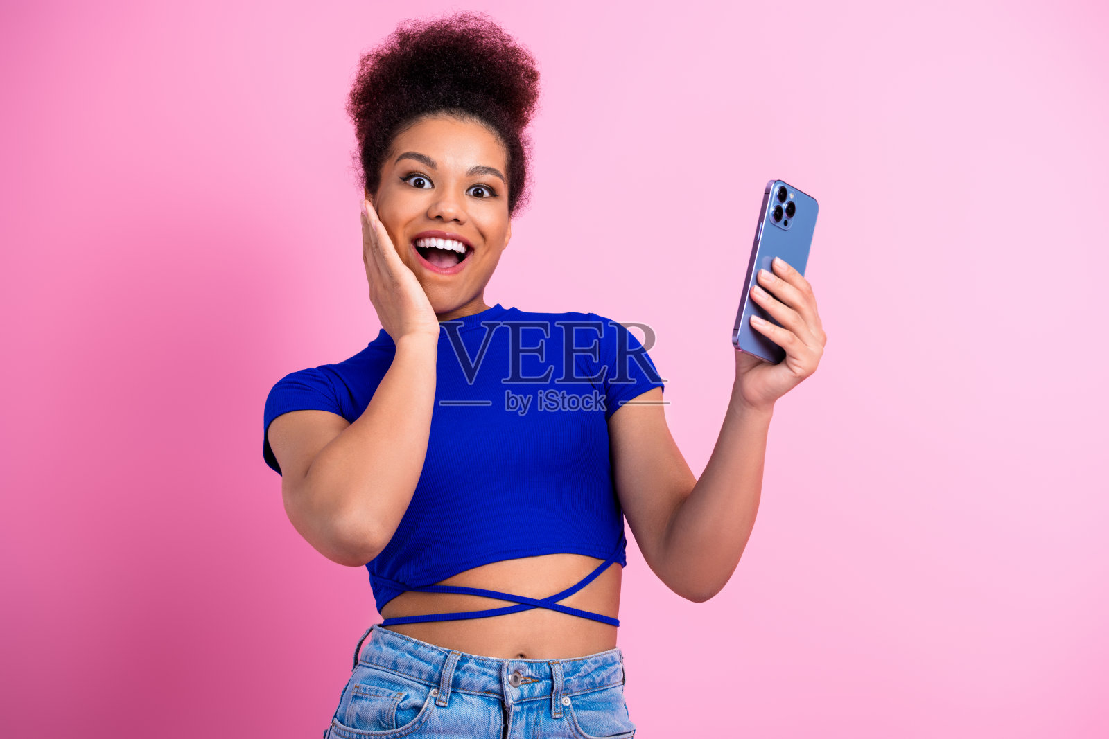穿着蓝色上衣的年轻女性手持智能手机，兴奋地在鲜艳的粉色背景前表现出喜悦。照片摄影图片