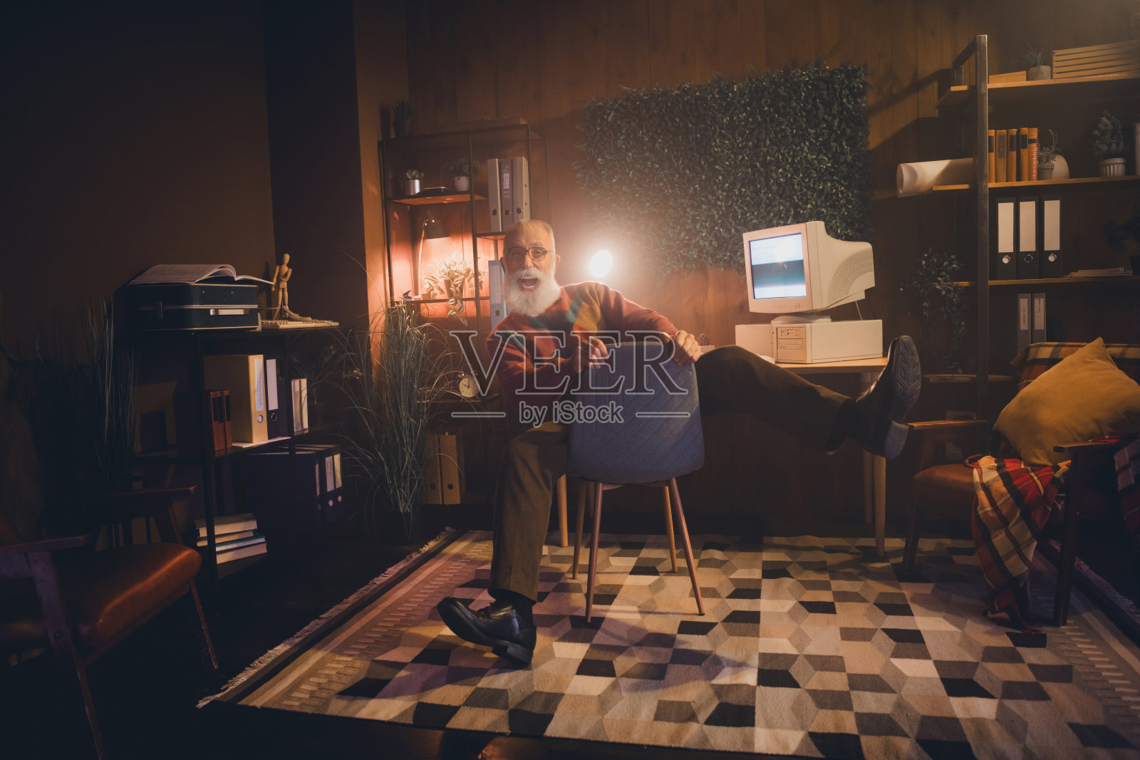 一位老年男子在温馨的办公室环境中使用复古电脑，周围有温暖的晚间氛围和复古风格的装饰。照片摄影图片