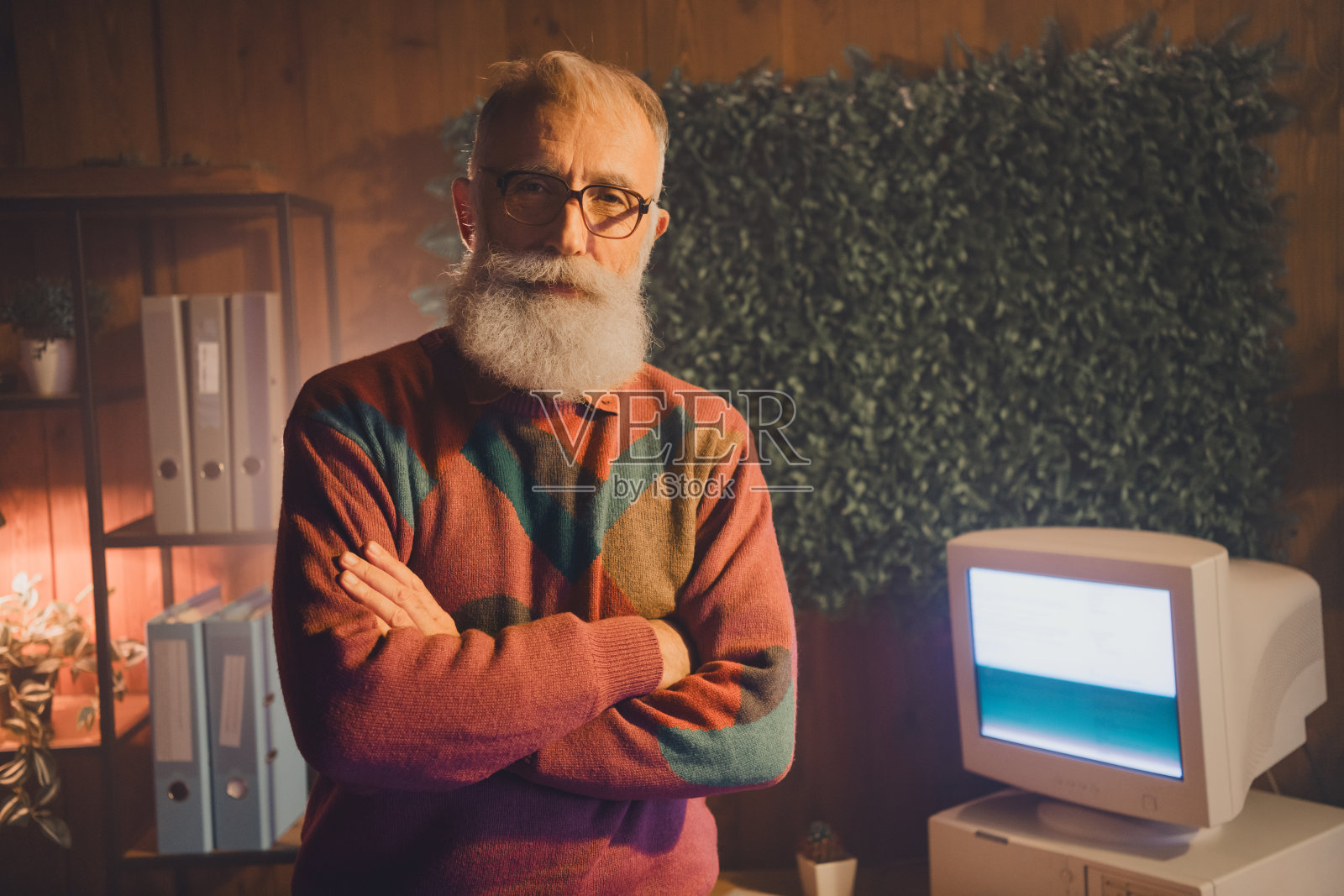 成熟男士穿着舒适的毛衣，自信地站在办公室里，身旁有一台复古电脑，展现出专业而放松的氛围。照片摄影图片