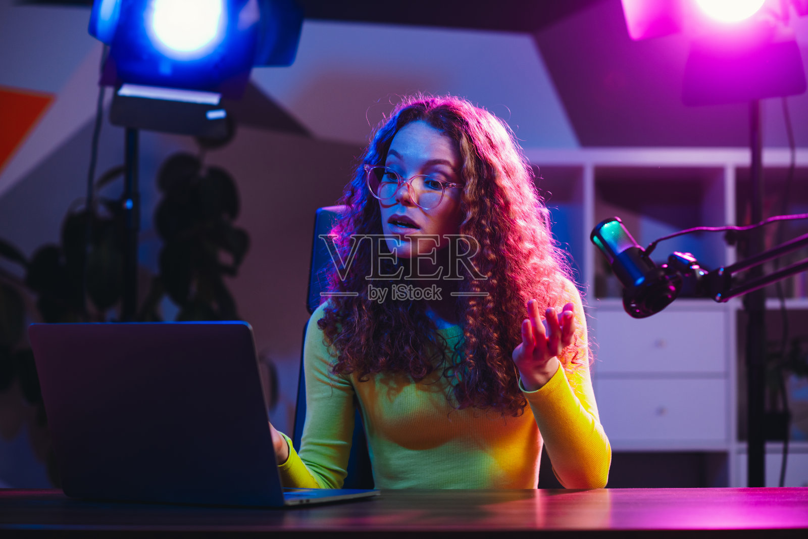 年轻女性在现代室内环境中使用笔记本电脑和麦克风进行直播，周围有明亮的霓虹灯。照片摄影图片