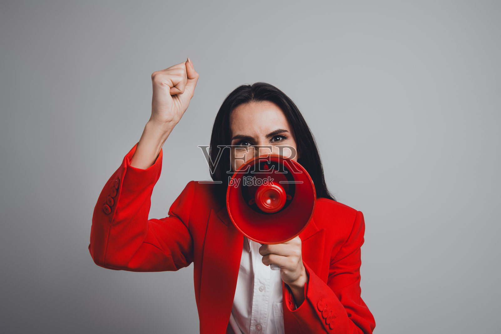 自信的女商人在灰色工作室中穿着红色西装，手持扩音器并举起手。照片摄影图片