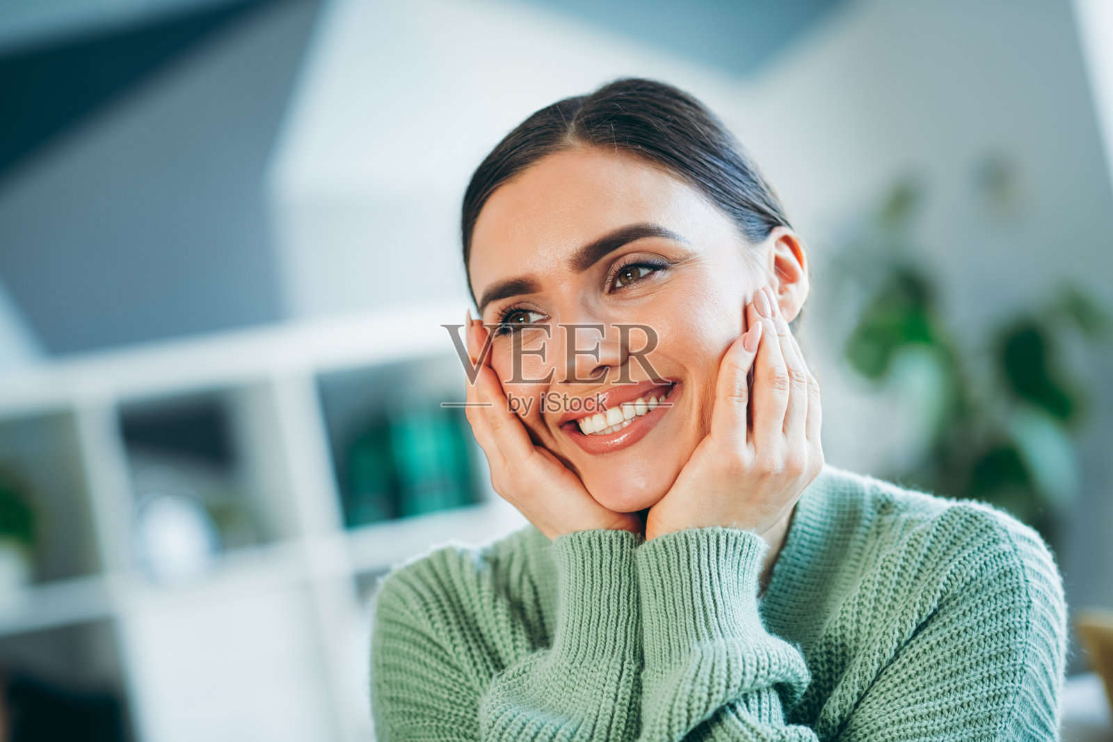 微笑的年轻女性双手托腮，享受着室内温馨的时刻，穿着绿色毛衣，身处明亮的家中。照片摄影图片