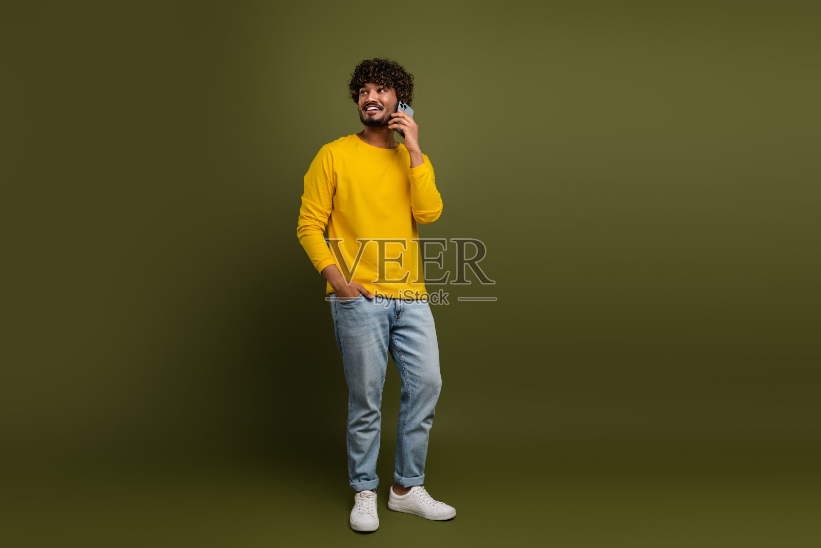 年轻男子穿着黄色毛衣，微笑着一边打电话，一边把手插在口袋里，背景为卡其色。照片摄影图片