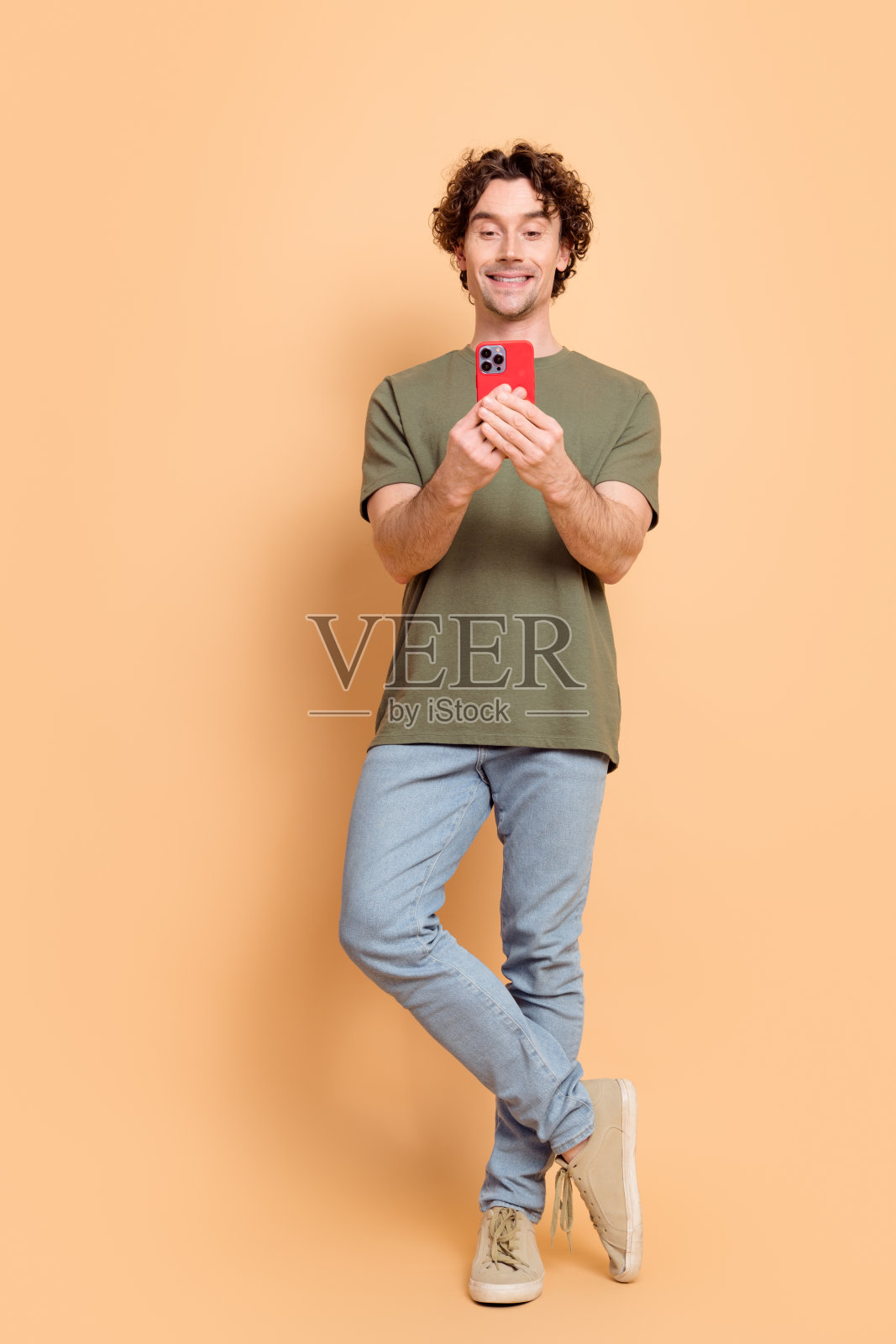 年轻男子穿着休闲卡其色衬衫，手持智能手机，面带愉快表情，时尚外观，背景为米色。照片摄影图片