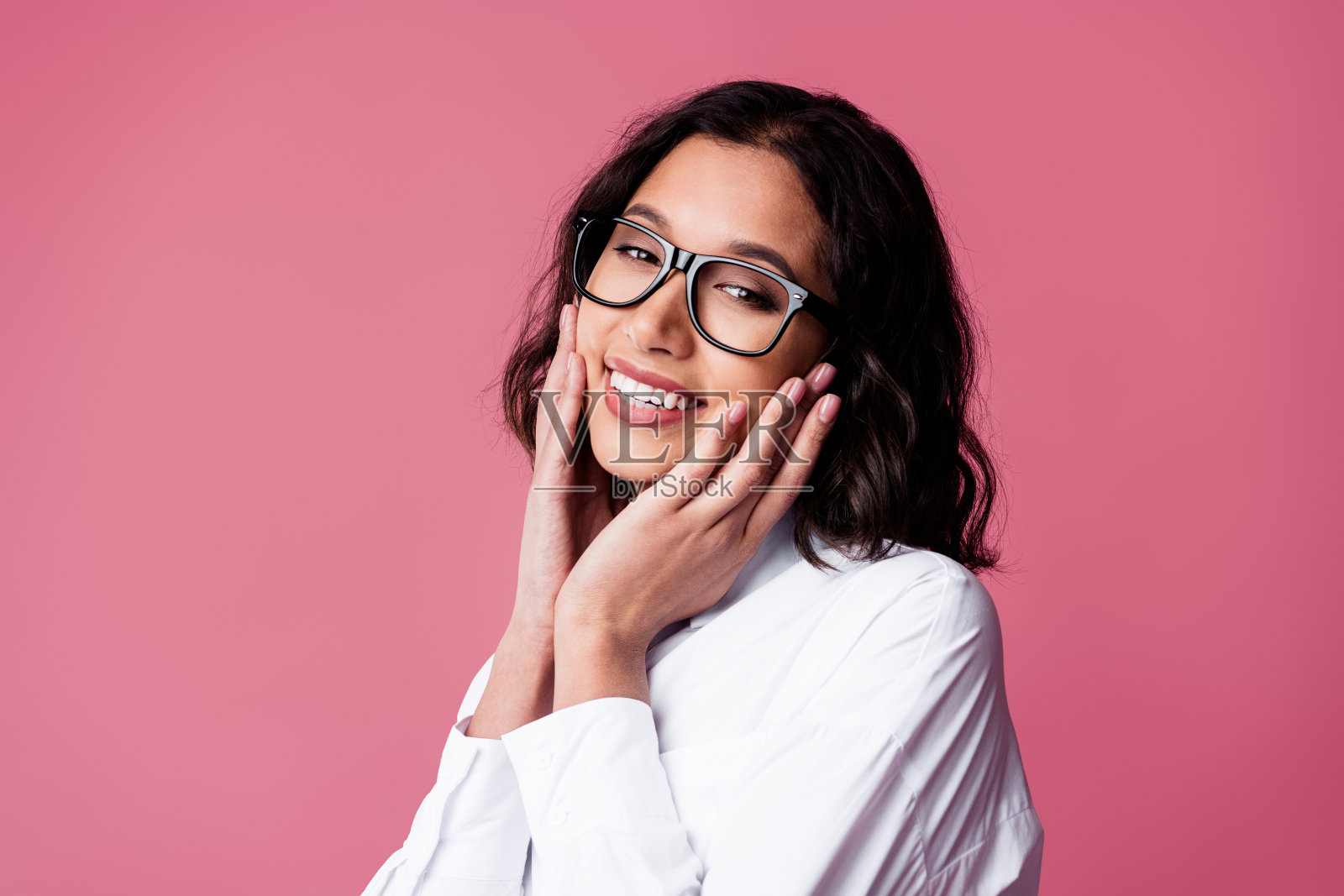 微笑的女性戴着眼镜，穿着时尚的白衬衫，自信地在粉色背景前摆姿势照片摄影图片